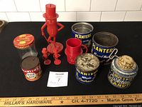 Overall lot showing Mr. Peanut bank, mugs, salt & pepper shakers and six meat and peanut tins placed on black cloth with measuring stick in front.