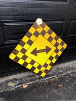 Photo of one diamond-shaped metal road sign with yellow and black checkerboard border featuring black arrows pointing left and right on a yellow background in the center.