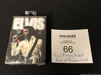 Sealed Elvis Presley playing cards with an image of Elvis in a white jumpsuit on front of box alongside auction lot number handwritten on paper.