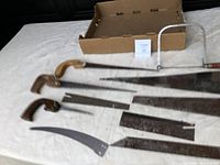 Slightly closer and angled view of the three wooden-handled hand saws and multiple loose saw blades on a white surface, showing details of handles and blade condition.