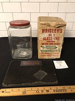 Complete lot showing glass jar, original box, and change mat on black surface with measuring ruler for scale.