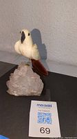 Photo of a white parrot figurine with reddish-brown tail perched on a clear quartz crystal cluster base, displayed on a flat surface with plain wall background.