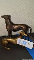 Two brass greyhound figurines positioned on a dark surface, one standing and one reclining, showing size and style differences.