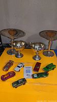 Silver compotes, sugar and creamer arranged behind assorted toy cars on yellow surface