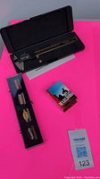Digital caliper in open black case with operating instructions, Craftsman pins in plastic display case, Marlboro lighter in cardboard sleeve on bright pink background