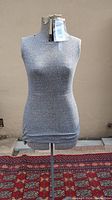 Gray dress mannequin torso on a metal stand against a beige wall and red patterned rug beneath (rug not included).