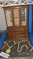 Wooden jewelry box with glass display case and three drawers, with necklaces hanging on the front and some jewelry pieces inside the case.