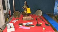 Full view of lot items on red surface showing bronze musician sculpture, brass peacocks, silverware, harmonica, pipe, and metal case
