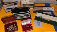 Overview of pens including Reflections, Cross, Omni, Taiwan Pen; gold-tone wristwatch; purple bead jewelry; and lapel pins in boxes.