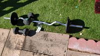 Photo showing two black 12 lb hex dumbbells and a chrome curl bar with four weight plates (two 5 lb and two 10 lb) placed on a grass surface and adjacent paving stones.
