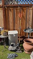 Outdoor yard items including Rubbermaid container, bird bath, terracotta pot, fruit picker, concrete statues, metal table, and chains arranged in sunlight.