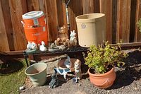Photo shows an assortment of garden statues, plant pots, and a large orange Igloo cooler against a wooden fence.
