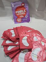 Photo showing the purple resealable bag with 120 pimple patches and a large pile of individually wrapped pink hydrogel eye patches.