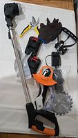 Photo showing electric lawn mower shaft with orange and black handle, pruning shears, protective gloves, two black and red batteries, charger, and interchangeable blades including circular saw and trimmer guard