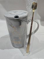 Photo of the marbled grey ceramic coffee cup with gold Sagittarius constellation design and lid, accompanied by gold-toned spoon still in plastic.