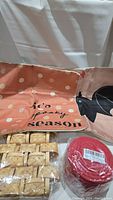 Photo shows two fabric pillow covers: a peach one with white polka dots and 'tis spooky season text, and a beige one with a black cat design. Six small woven baskets and a plastic wrapped set of red coasters in front.
