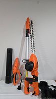 Image showing orange cordless hedge trimmer with extendable pole and black handle, black battery charger, and carrying case.