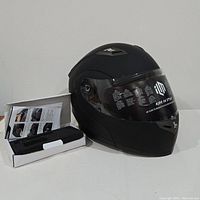 Matte black ILM motorcycle helmet with Bluetooth module shown next to box of accessories, visor down with protective film attached.