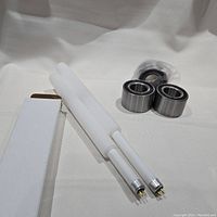 Two new fluorescent light tubes with metal bases beside four metal bearings, two of which are sealed in plastic bags.