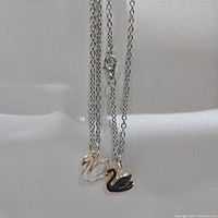 Two necklaces with swan pendants, white and black variants, displayed hanging against a light fabric background.