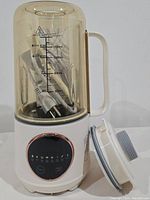 Front view of soy milk maker showing amber tinted container with handle and control panel on white base. Lid placed beside.