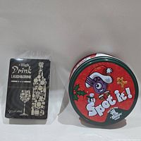Photo showing Spot It! game in its festive red round tin and Laugh And Drink card game in sealed black packaging.