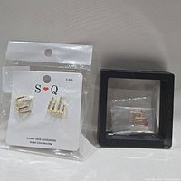 Two pairs of earrings. One pair gold-tone with rhinestones on S Q card and packaged in plastic; the other pair rose gold-tone square hoop earrings in a small black frame box.