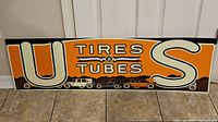 Front view of rectangular metal sign with 'U S Tires & Tubes' graphics in orange, white, and brown, vintage car silhouettes along bottom