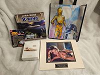 Full view of the lot showing the Star Wars X-Wing board game, Star Wars Metal Earth Millennium Falcon model kit box, C-3PO and R2-D2 poster tote, Princess Leia framed art, and Star Wars cards.