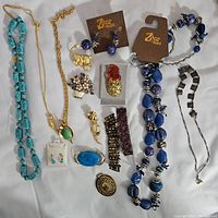 Top-down photo showing an assortment of costume jewelry including turquoise and blue necklaces, earrings on Zbar cards, several brooches, rings, and bracelets laid out on white background.