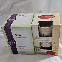 Photo of Scentsy Stripe plug-in wax warmer in original green and purple box and Candle Warmers Live Laugh Love pluggable fragrance warmer in cream box with window showing warmer