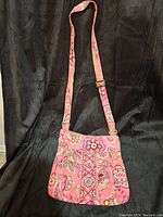 Pink Vera Bradley crossbody bag with adjustable strap hanging against dark background