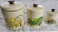 Full view of three metal canisters of varying sizes with nature-themed painted decoration and brown knob lids