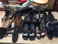 Lot of 15 pairs assorted shoes including high heels, tall boots, sandals, moccasins, sneakers, Puma sandals, various styles and colors on a table