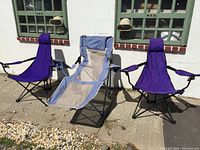 Photo showing the three foldable camping chairs outside against a white wall and green window frames, two purple and one blue/tan reclining chair arranged side by side.
