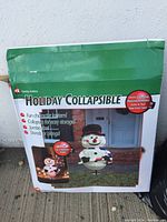 Box showing the product name and image of the collapsible snowman lantern in a front porch setting.