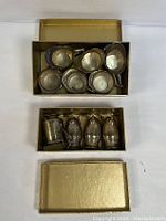 Top view of two open gold-colored boxes containing six small metal pitchers and vases with metallic silver finish.