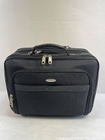 Front view of black Samsonite travel bag showing two main zippered compartments, front pocket with metal brand plate, and top handle