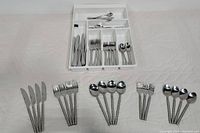 Overall view of two flatware sets in white plastic tray and arranged on white cloth surface showing knives, forks, and spoons with twisted handle design and second set in tray with simple handles.