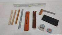 Photo shows a variety of measuring tools arranged on a white surface, including wooden and metal rulers, leather cases, tape measures, a small folding square, and a refrigerator thermometer.
