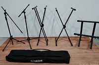 View of seven black metal stands including two microphone stands with boom arms and four X-style keyboard or instrument stands beside a black On-Stage Stands carry bag.