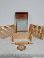 Front view of the vintage National Washboard No. 512 with metal scrubbing area and wooden frame, flanked by two shallow slatted wood crates, a wooden wall shelf in front, a traditional wooden rolling pin, and a circular wooden butter dish base.