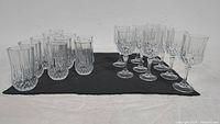 Two groupings of glasses displayed on a dark mat. Left side shows 10 diamond cut pattern highball glasses. Right side shows 10 stemmed wine glasses with cut detailing on bowl and textured stems.