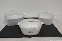Three white ceramic casserole dishes with clear glass lids shown from the front on a black mat, one Corning Ware with black floral design and two plain white dishes.