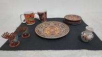 Front view of the Imperial Fine China plate and saucers, two Kokopelli mugs, Hopi pottery jar, and two Kokopelli candleholders on a dark mat.