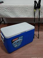 Blue Coleman cooler with white lid and label showing 45.4 liters capacity next to a pair of black trekking poles standing vertically.