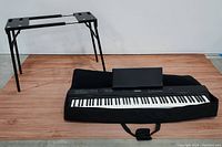 Full view of the Priva electronic keyboard laid on the black soft carrying case with music stand attachment on keyboard, and black metal adjustable keyboard stand next to it.