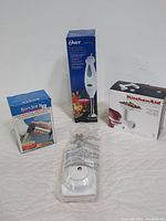 Four kitchen appliance items: Oster hand blender in box, Bonjour hand torch box, Pampered Chef Simple Slicer box, KitchenAid food grinder attachment box.
