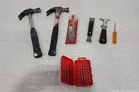 Full view of all tools on white quilted surface including two hammers, socket set, drill bits in case, scrapers, and screwdriver.