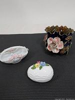 Three vintage flower-themed ceramic items on black surface: a dark glazed vase with large pink roses, a white oval dish with small pink roses, and a white egg-shaped trinket box with colorful small flowers on lid.